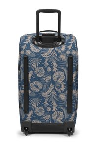 Rullattava matkalaukku sinisellä taustalla, jossa on beigejä trooppisia lehtikuvioita. Kova kuori, säädettävä kahva ja mustat yksityiskohdat.