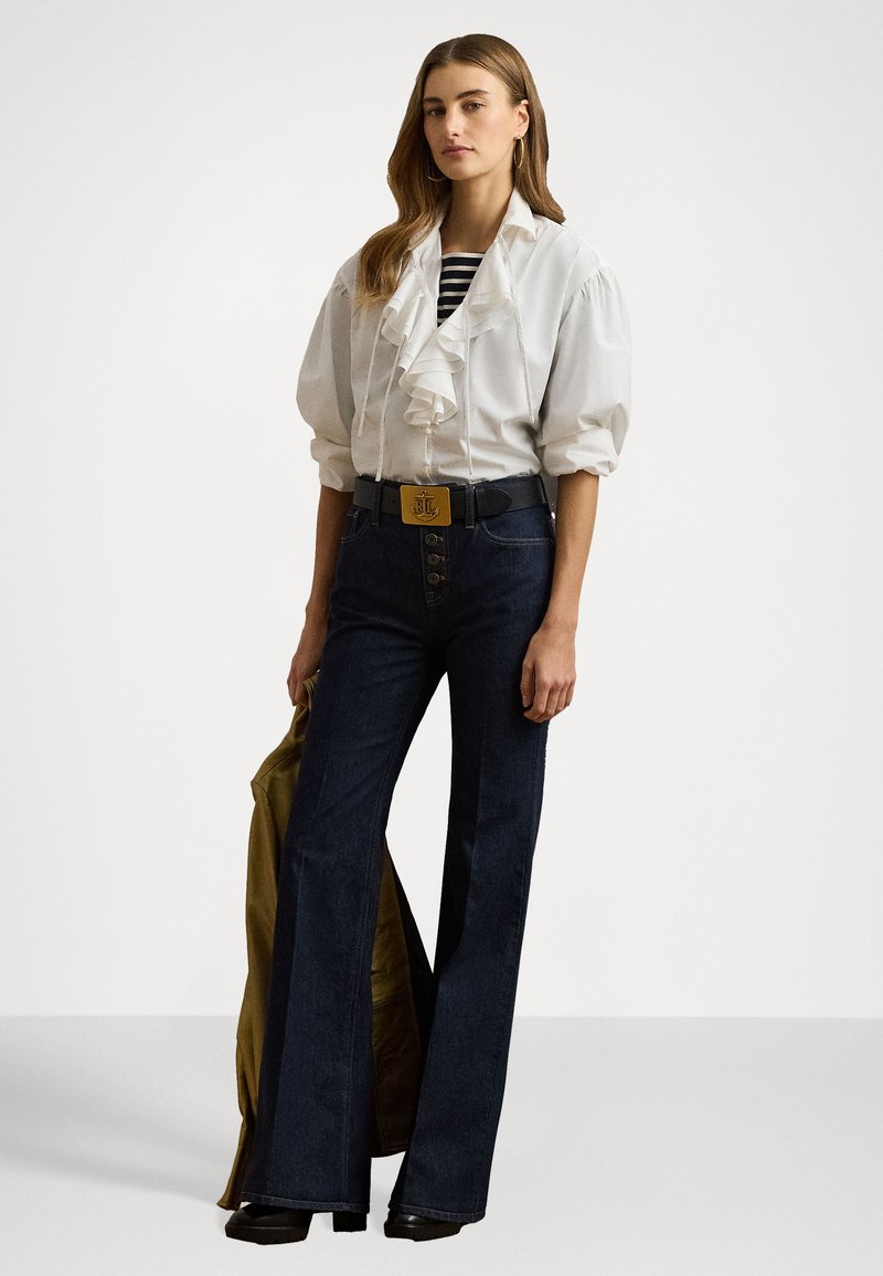 Biała bluzka z falbanami i obszernymi rękawami, w paski podkoszulek, ciemnoniebieskie dzwony, czarny pasek z złotą klamrą, stojąca na jednolitym tle.