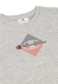 Camiseta de algodón gris con cuello redondo, que presenta un logo en forma de diamante en negro y rojo, con patrones de rayas horizontales.