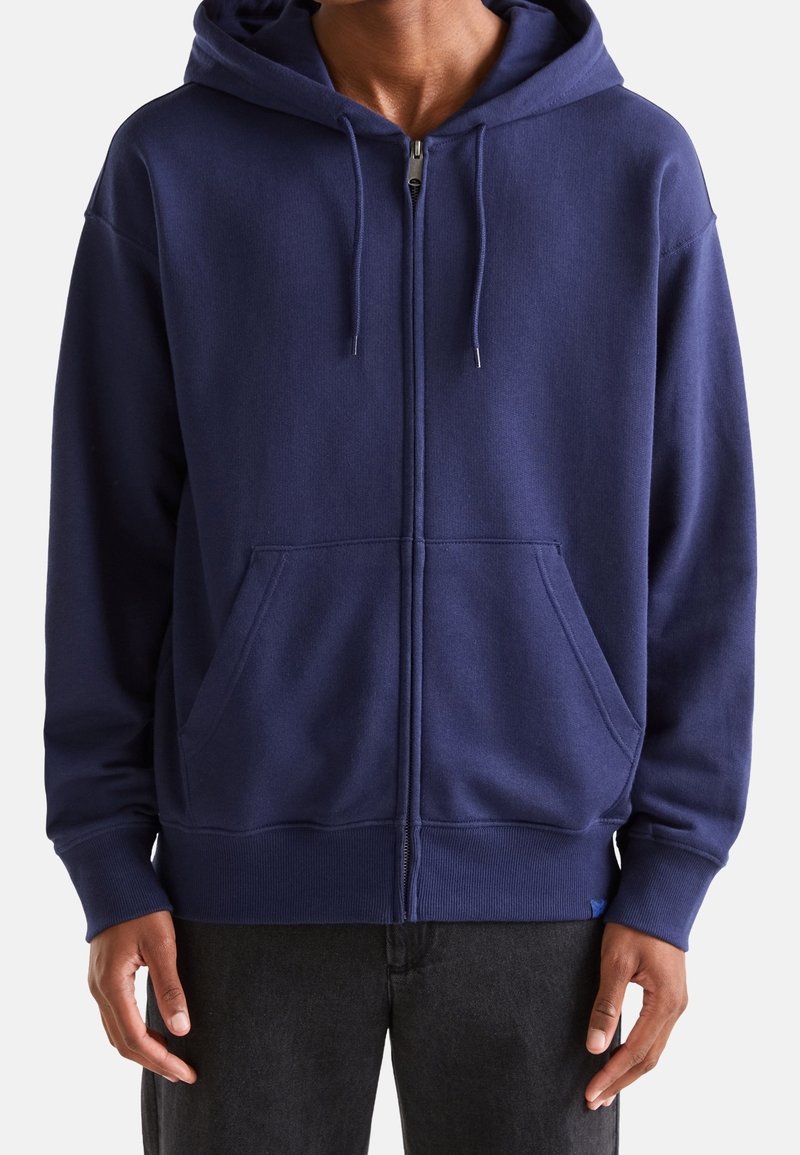 Muž oblečený v tmavě modré mikině na zip s předními kapsami a šňůrkami, v kombinaci s černými kalhotami, stojící na bílém pozadí.