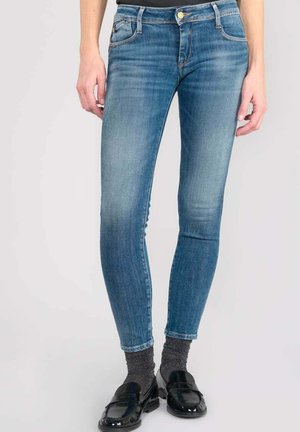 Jean skinny bleu, taille mi-haute avec un délavage, cinq poches, accent de bouton doré, tissu en denim extensible, associés à des mocassins noirs et des chaussettes grises.
