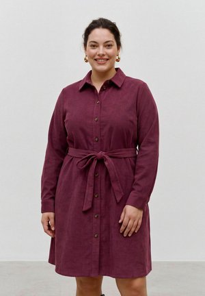 Femme aux cheveux foncés portant une robe bordeaux boutonnée avec une ceinture à la taille, debout et souriante sur un fond clair uni.