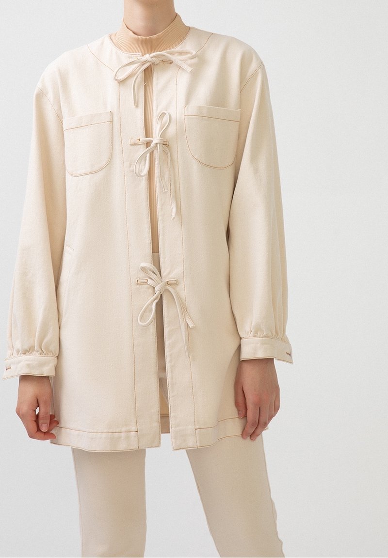 Veste en coton beige avec des manches longues, deux poches avant, des liens réglables au niveau du cou, et un col et des poignets côtelés. Design minimaliste.