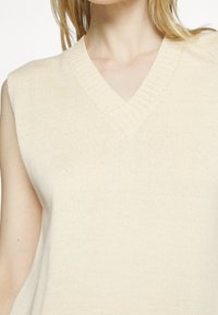 Pull sans manches beige avec un décolleté en V, fabriqué en tissu tricoté texturé, comportant des détails côtelés le long de l'encolure.