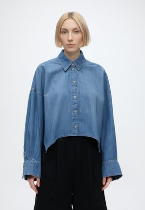Jonge vrouw met kort blond haar die een oversized cropped denim blouse met knopen en losse zwarte broek draagt, staand tegen een effen achtergrond.
