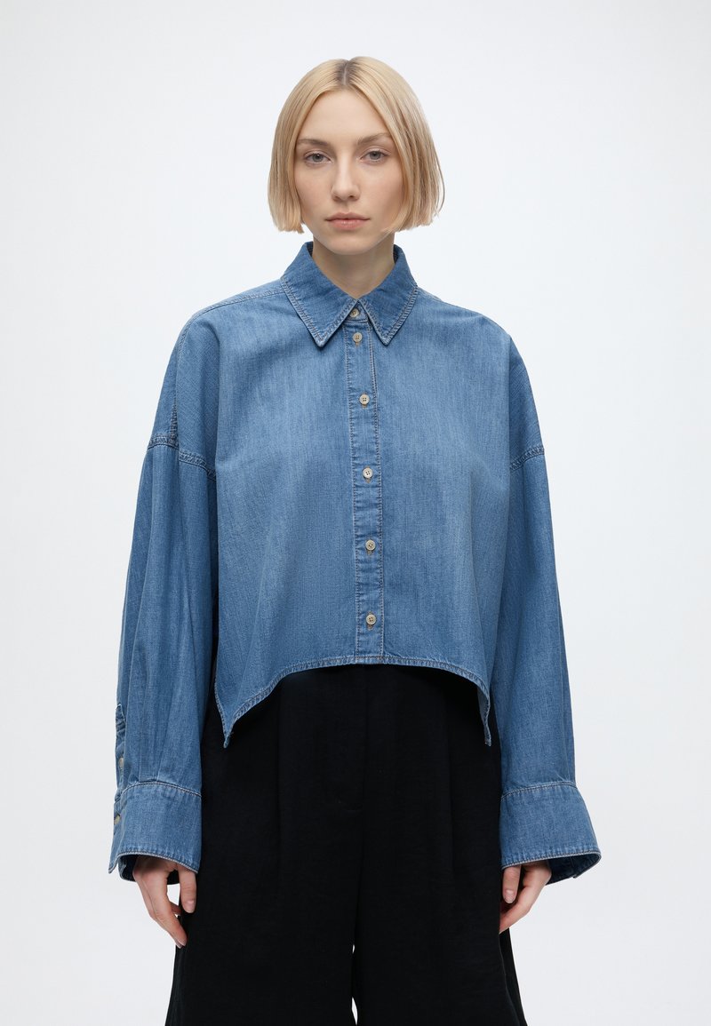 Jeune femme aux cheveux blonds courts portant une chemise en jean oversize coupée courte avec des boutons et un pantalon noir ample, se tenant devant un fond uni.
