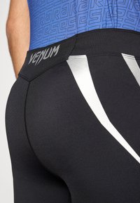 Leggings atléticas pretas com textura suave, apresentando detalhes prateados e um logótipo "VENUM" em negrito na cintura. O top é azul com um padrão geométrico.