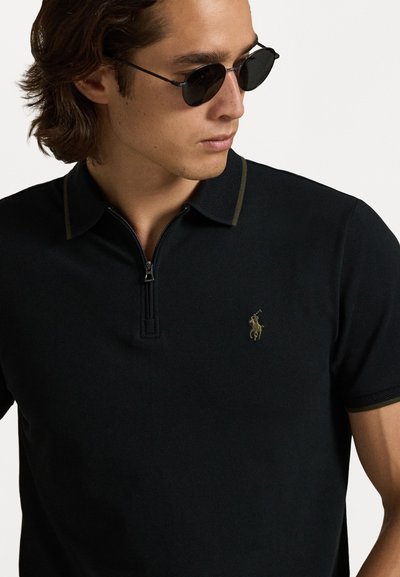 Svart polo skjorte med korte ermer, glidelås krage og kontrasterende grønne detaljer. Har brodert logo på brystet. Vevd tekstur.