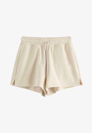 Kvinde står iført løs beige hættetrøje, matchende shorts, hvide sokker og hvide sneakers mod en ensfarvet lys baggrund.