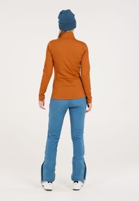 Oranje lange mouwen top en blauwe skibroek, met een relaxte pasvorm en zachte textuur. De broek heeft ritsdetails bij de enkels.