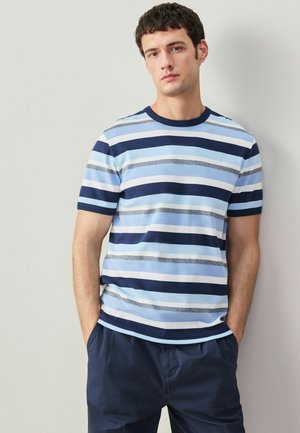 Junger Mann steht mit Händen in den Taschen und trägt ein kurzärmliges, gestreiftes Hemd in Blau- und Marineblau-Tönen sowie eine marineblaue Hose.