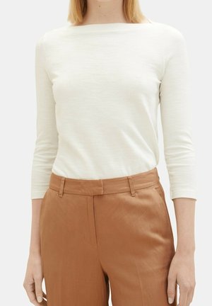 Mujer con una camiseta blanca de manga larga metida dentro de unos pantalones de tiro alto color marrón claro con trabillas para cinturón y bolsillos laterales.