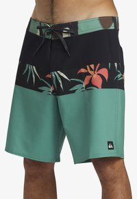 Bañador tricolor con cinturilla de marinero, sección inferior verde y patrón floral negro en la parte superior. Cuenta con cordones en la parte delantera y parche con el logo.