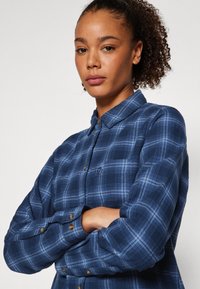 Camicia a quadri blu navy con colletto button-down, maniche lunghe e dettagli in bottone dorato. Presenta un logo ricamato sottile sul petto.