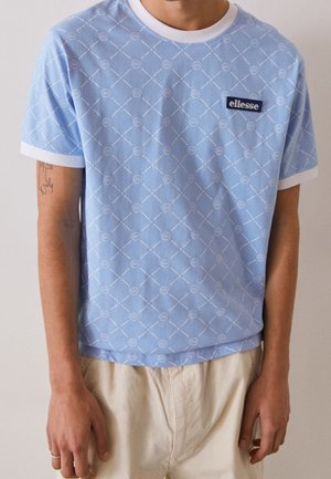 Homme portant un t-shirt light blue ellesse avec un imprimé blanc à motifs et des bordures blanches sur les manches, associé à un pantalon beige.
