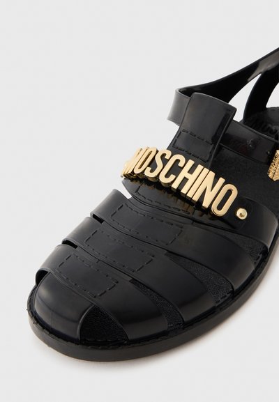 Must kivistatud kummijalatsid, millel on kinnine varvas ja avatud konts. Ees on kullavärvi "MOSCHINO" logo ning tekstuuritud triibud.