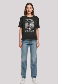 Black cotton t-shirt featuring a graphic print with "JUSTIN" and "BIEBER" text in white, paired with light blue denim jeans and white sneakers.