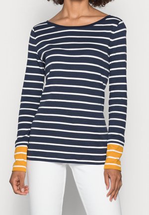 Vrouw die een marineblauw-wit gestreept top met lange mouwen en gele manchetten draagt, gecombineerd met een witte broek, staand tegen een effen achtergrond.