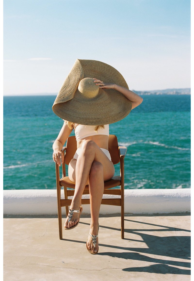 Cappello di paglia a tesa larga, costume da bagno bianco, pelle abbronzata, seduto su una sedia di legno, gambe incrociate; sandali decorati con accenti ornamentali.