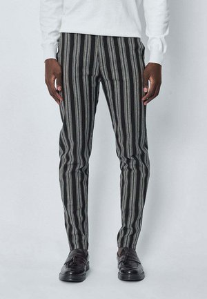 Homme portant un pantalon slim rayé verticalement noir et gris, un pull blanc, et des mocassins en cuir noirs à gland, debout avec les mains relaxées.