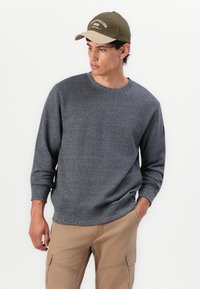 Felpa grigia a maglia con scollo rotondo e polsini a coste, abbinata a pantaloni cargo color khaki chiaro e un cappellino bicolore con scritta.
