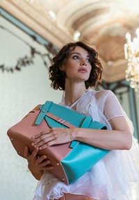 Žena ve průhledné bílé blůze drží velkou dvoubarevnou strukturovanou kabelku modré a růžové barvy v elegantní místnosti s lustr.