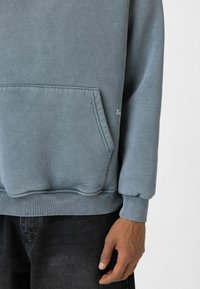 Pullover hoodie in zachte blauwe tint, gemaakt van een zachte stof. Heeft een kangoeroezak, ribgebreide manchetten en onderrand. Minimalistisch geborduurd logo-detail.
