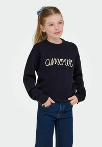 Dekleta s čopom, oblečena v mornarsko modro jopico s textom "amour" in modre kavbojke, stoji z rokama v žepih pred svetlo ozadje.