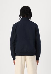 Veste bleu marine avec col, poignets élastiques et taille élastique ; présente un design de panneau arrière et une texture lisse. Associée à un pantalon de couleur claire.