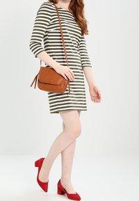 Robe rayée vert olive et blanc, sac bandoulière marron, chaussures à talons carrés rouges, et teint de peau pâle. Design simple avec des manches aux trois quarts.