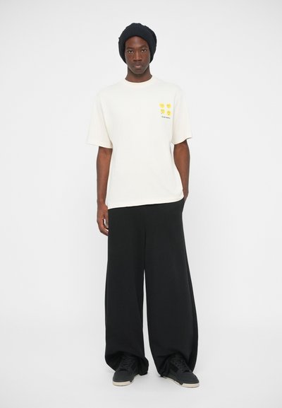 White t-shirt with yellow graphic design and text on the chest, paired with loose black pants and black sneakers. Black knitted beanie worn.