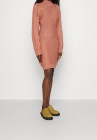 Robe tricotée couleur rose avec un col haut et des manches longues, associée à des sabots jaunes avec une semelle noire. Texture lisse et design ajusté.
