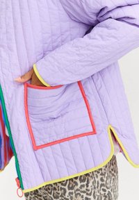 Dłoń włożona w kieszeń lawendowej pikowanej kurtki z czerwonym wykończeniem, żółtymi mankietami oraz zamkiem błyskawicznym w zielonym i niebieskim kolorze, noszonej na spodniach w panterkę.