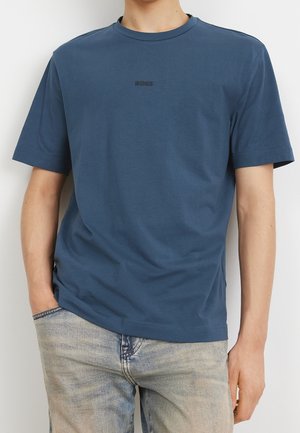 Mann mit einem schlichten blauen Kurzarm-T-Shirt mit "BOSS"-Textlogo auf der Brust und ausgewaschenen hellblauen Jeans, eine Hand in der Tasche.