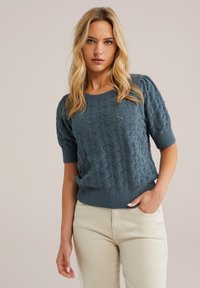 Maglione a maglia di un azzurro chiaro con maniche a palloncino corte, caratterizzato da un motivo a diamante. L'orlo e i polsini a coste completano il design. Indossato con pantaloni crema.