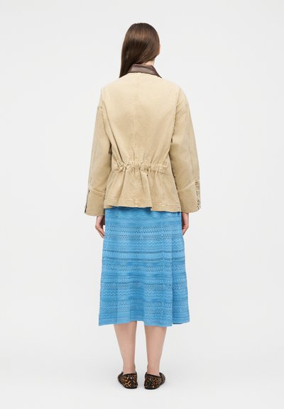 Femme aux longs cheveux bruns portant une veste beige à taille cintrée par un cordon, une jupe midi texturée bleue et des ballerines imprimé léopard, vue de dos.