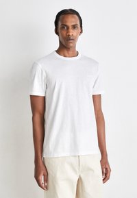 Weißes Baumwoll-T-Shirt mit Rundhalsausschnitt und kurzen Ärmeln, das ein kleines Logo auf der Brust trägt. Glatte Textur, lockere Passform und klassisches Design.