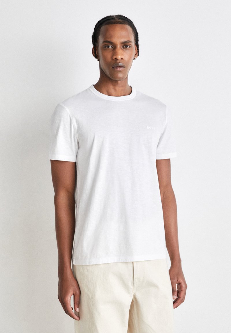 Weißes Baumwoll-T-Shirt mit Rundhalsausschnitt und kurzen Ärmeln, das ein kleines Logo auf der Brust trägt. Glatte Textur, lockere Passform und klassisches Design.