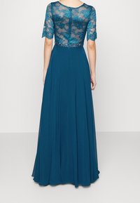 Femme portant une robe bleu sarcelle longue jusqu'au sol avec un dos et des manches en dentelle transparente, une fermeture éclair et une ceinture ornée de perles.