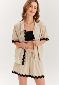 Béžová lněná košile s černým okrajem, doplněná o odpovídající šortky se stejným okrajem. Košile s knoflíky a elastickým pasem.