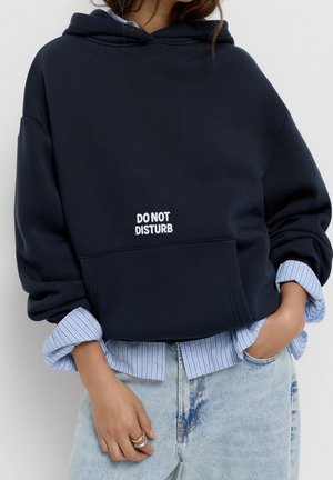 Person trägt einen marineblauen Hoodie mit dem Text "DO NOT DISTURB", über einem hellblau gestreiften Hemd und hellblauen Jeans. Eine Hand in der Tasche.