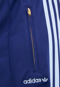 Jūrinės sportinės kelnės su šviesiai mėlynomis juostelėmis ir užtrauktuku ant kišenės, kuri turi auksinį užtrauktuką. Apatinėje kojoje yra balta logotipo žymė.