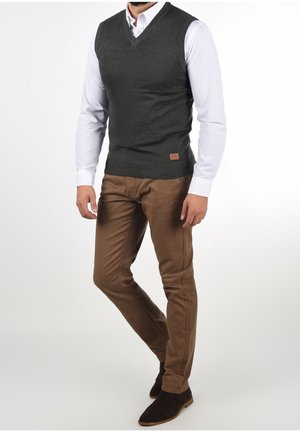 Gilet grigio a maglia sopra una camicia bianca, abbinato a pantaloni slim-fit marroni e stivali alla caviglia marroni scuri. Presenta una piccola piastrina in pelle sul gilet.