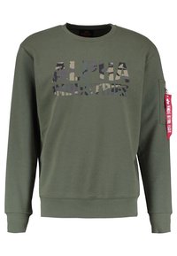 Felpa verde girocollo con testo in camo "ALPHA INDUSTRIES" sul davanti e un'etichetta rossa "Remove Before Flight" sulla tasca della manica sinistra.