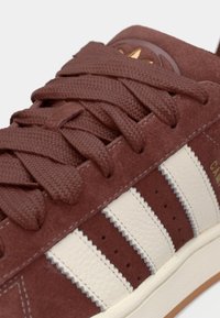 Brune ruskinds sneakers med hvide læderstriber, teksturerede snørebånd og et lille guld-accent på tungen. Gummisål er synlig.