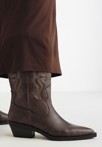 Botas western de piel marrón con puntas afiladas, costuras decorativas y un tacón resistente. Presenta detalles acentuados en un patrón ondulado.