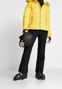 Gelbe Pufferjacke mit Fellbesatz an der Kapuze, schwarze Skihosen und schwarze Skischuhe. Hält einen schwarzen Skihelm mit klarem Visier.