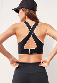 Zwarte sportbeha met kruisrugontwerp en witte afwerking. Beschikt over een driehoekige uitsnede aan de achterkant. Model draagt zwarte leggings met logo.