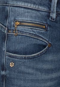 Jeans de mezclilla azul oscuro con un bolsillo con cremallera y herrajes dorados, costuras en contraste y un pequeño emblema metálico en el bolsillo delantero.