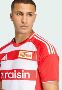 Červený a biely pruhovaný športový dres s logom, krátkymi rukávmi, okrúhlym výstrihom a tromi bielymi pruhmi na pleciach, vyrobený z priedušnej látky.